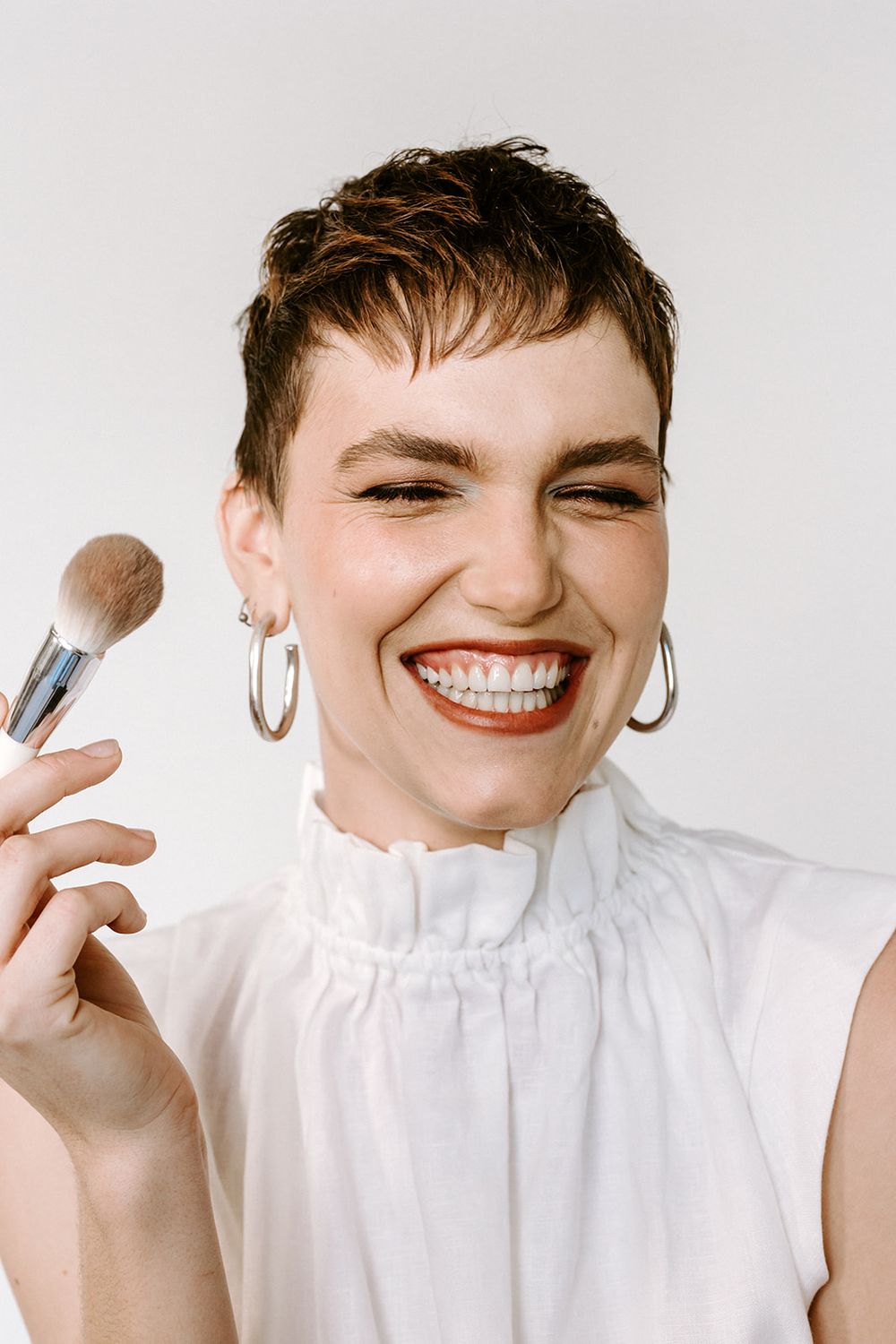 Smiling woman with short hair holds a makeup brush, wearing red lipstick, hoop earrings, and a white ruffled top.