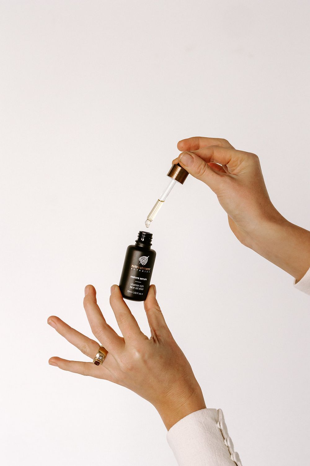 Hands holding a dark glass dropper bottle of facial serum against a white background, with a gold ring accent.