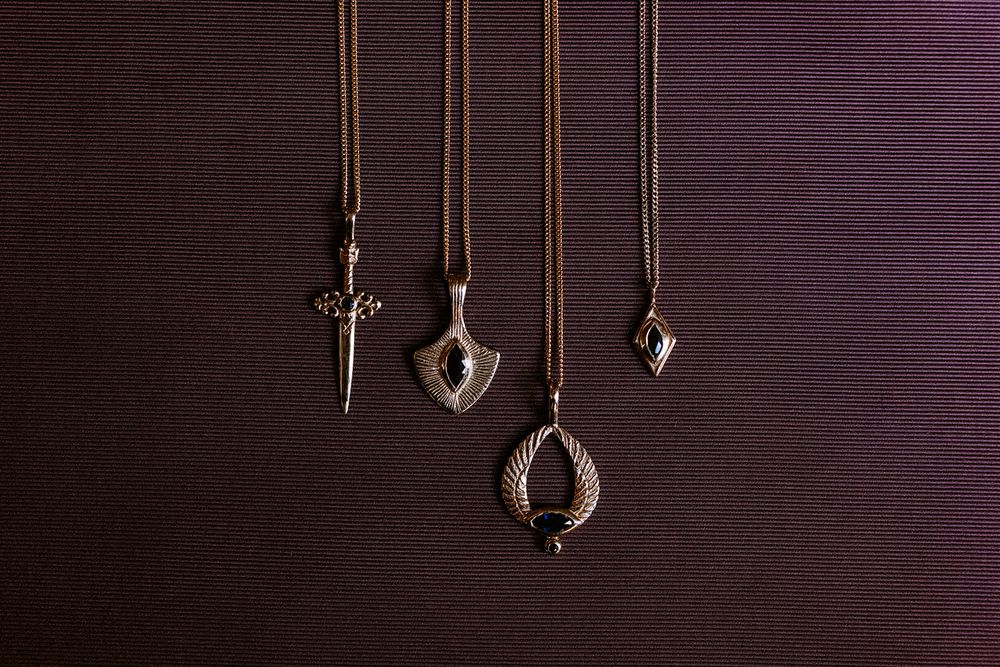 Four silver pendant necklaces on leather cords displayed against dark background, featuring sword, shield, teardrop, and diamond shapes.