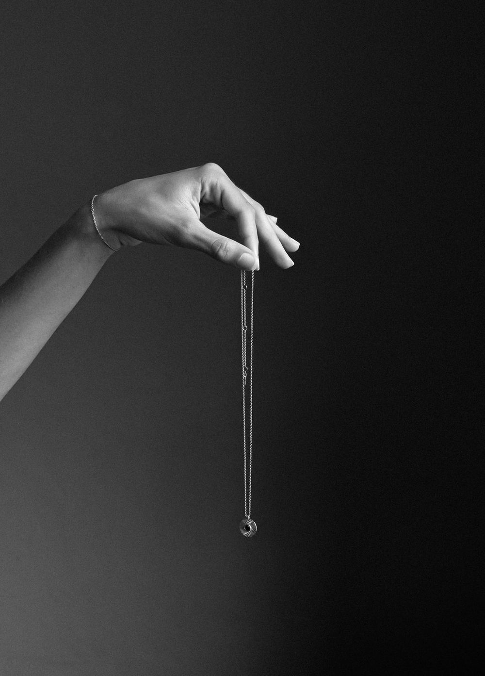 A hand delicately holds a thin silver necklace with a small pendant against a dark background in black and white.