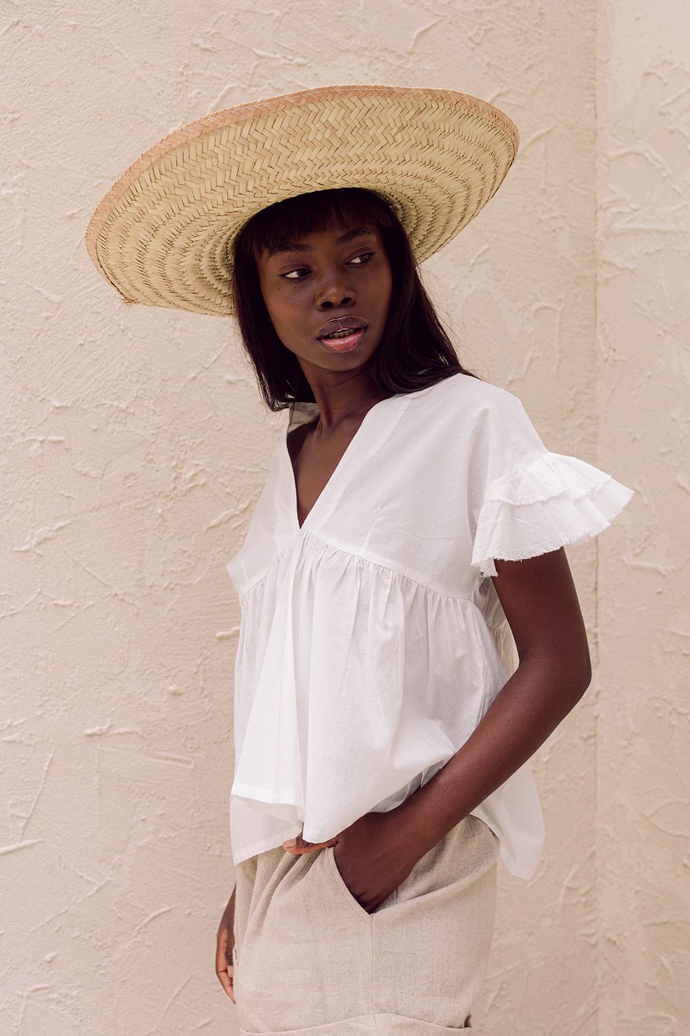 Woman in white ruffled blouse and wide-brim straw hat posing against a textured beige wall.