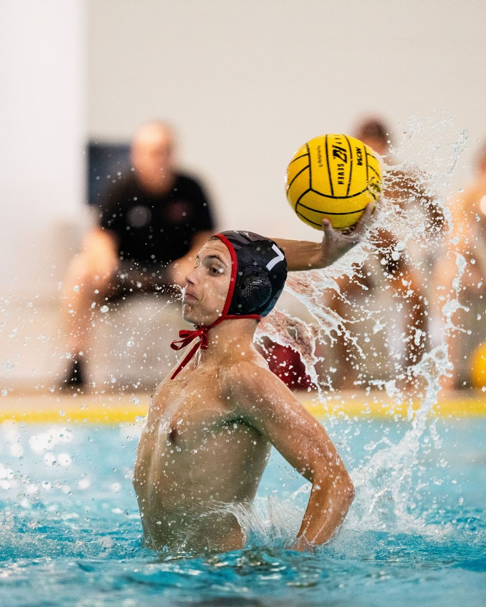 Water polo player rises from pool to throw yellow ball during competitive match indoors.