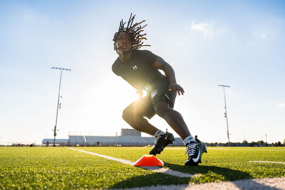 Athlete sprints past orange cone on green turf field with bright sunlight glowing behind them.
