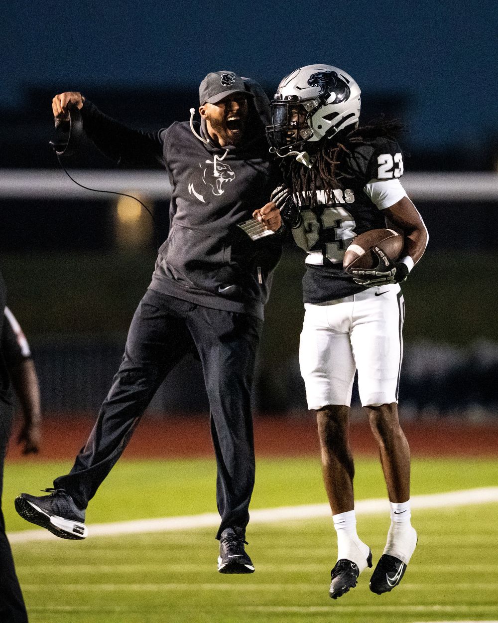 Football coach celebrates with player number 23 on the sideline during a night game.