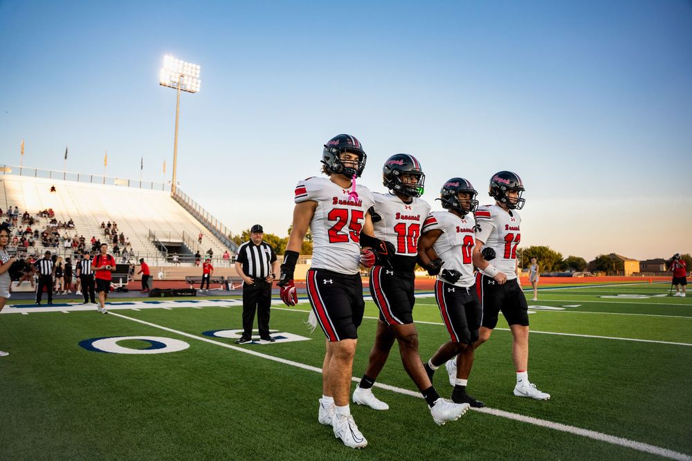 Four Braswell football players walk onto the field at dusk before a high school game.