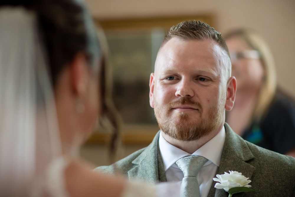 Groom in green tweed suit with boutonniere gazes lovingly at bride during wedding ceremony.
