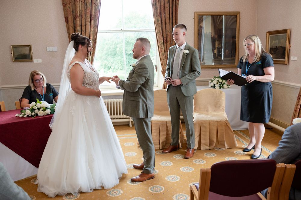 Bride and groom exchanging rings during wedding ceremony with officiant and guests in a formal venue.