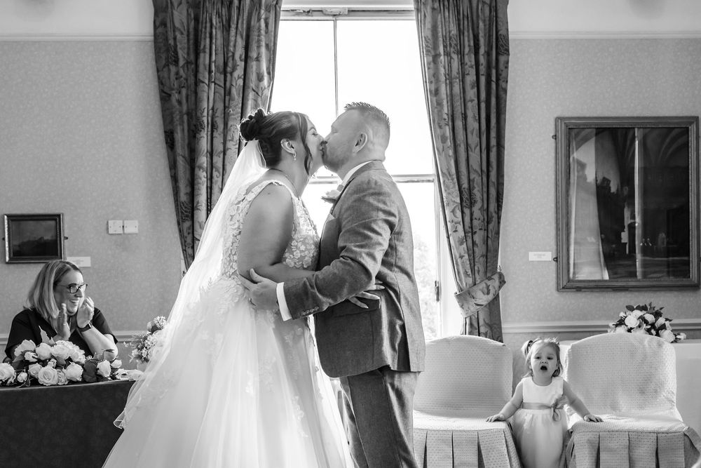 Black and white wedding photo of bride and groom sharing first kiss as flower girl reacts with delight in background.