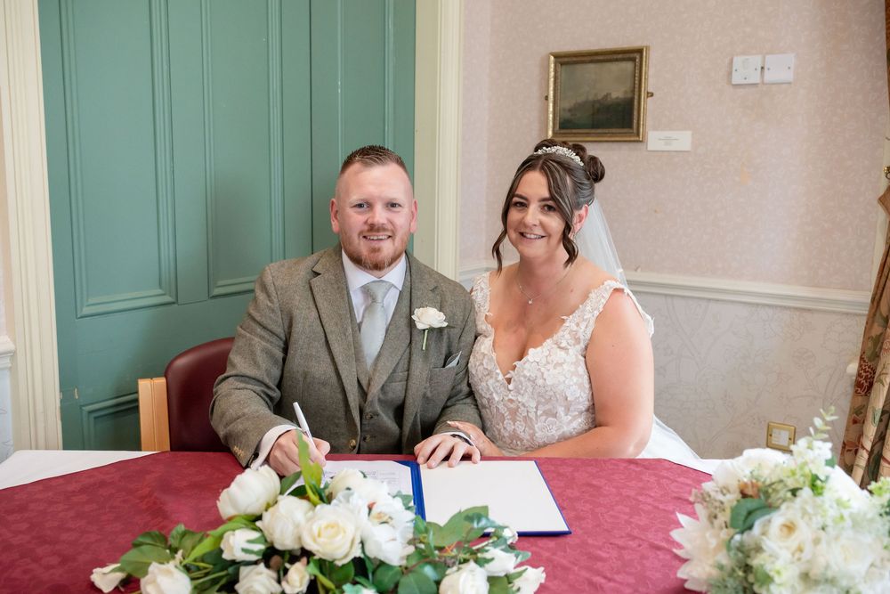 Smiling bride and groom signing wedding register at decorated table with white floral arrangement.
