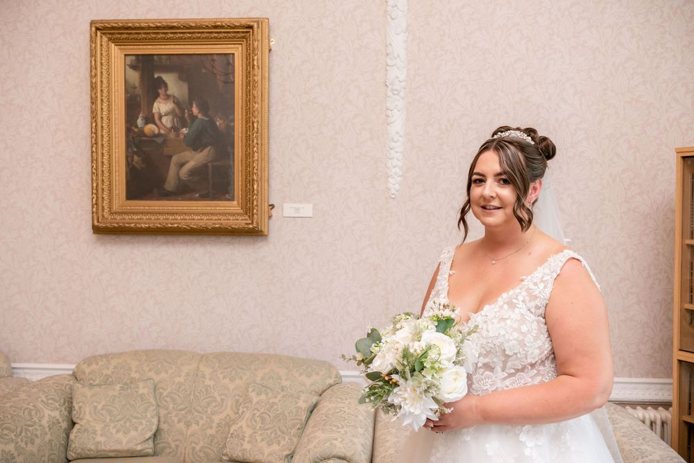 Bride in lace wedding dress holding white bouquet stands smiling beside vintage painting in elegant room.