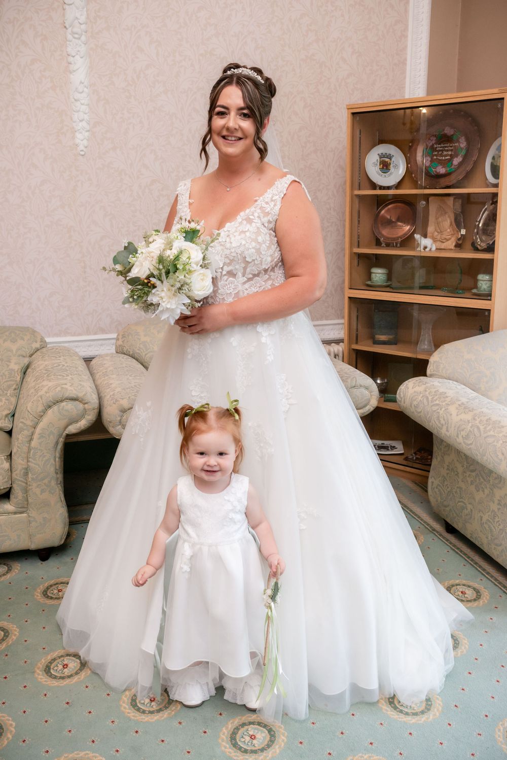 Bride in white lace ballgown holding bouquet poses with young flower girl in white dress indoors.