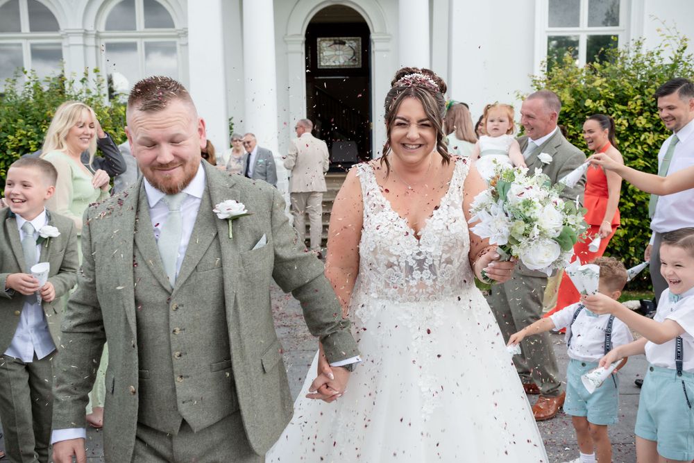 Bride and groom walk hand in hand through confetti outside a white church surrounded by wedding guests.