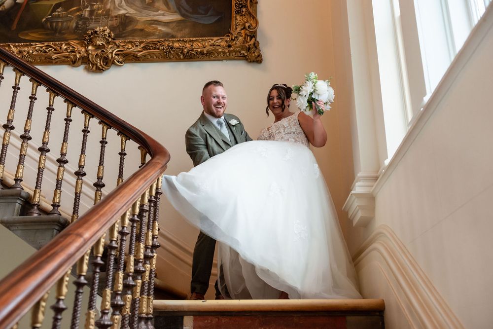Bride and groom descend elegant staircase, bride in white ballgown holding bouquet, groom in grey suit at wedding venue.