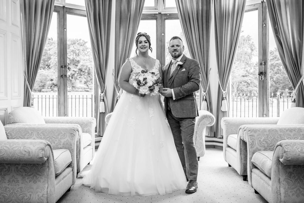Bride and groom posing together in elegant wedding venue with large windows and draped curtains in black and white photo.
