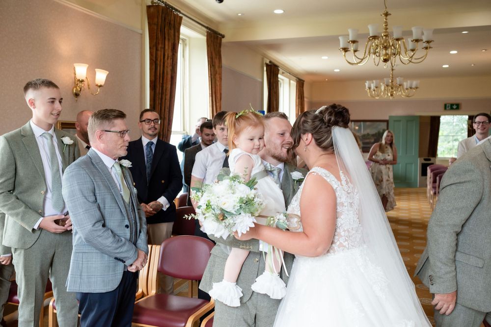 Bride in white lace gown kisses child at wedding ceremony while groomsmen stand nearby in elegant venue.