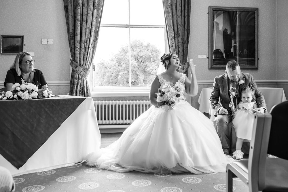 Black and white wedding photo of bride in ball gown seated with groom and guests during ceremony indoors.