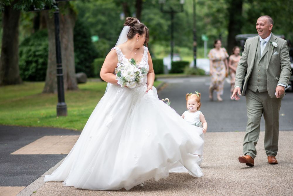 Bride in white gown holds bouquet with flower girl on path, groom and bridesmaids following in lush green park.