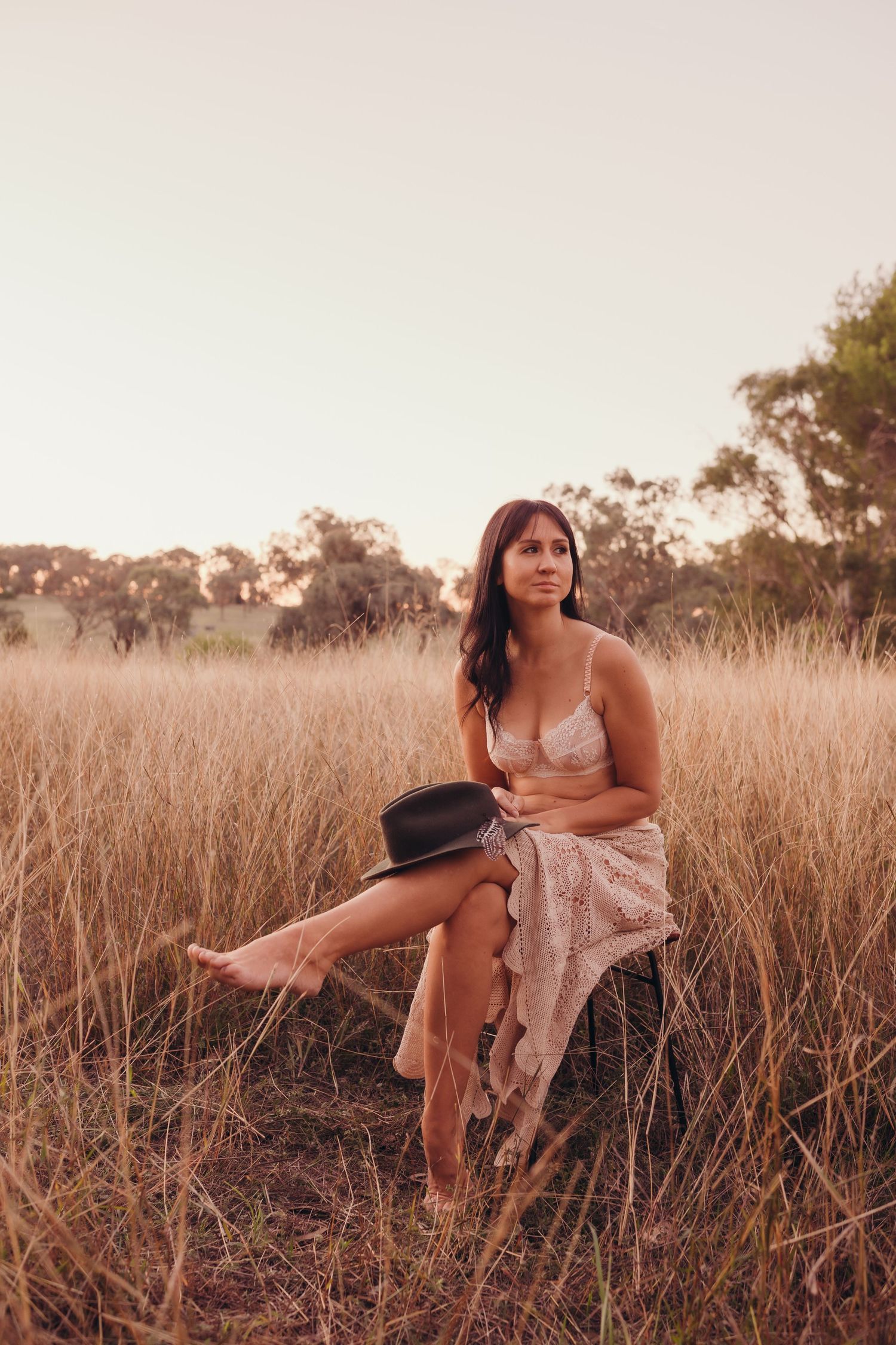 Young woman in lace lingerie and skirt sitting in dry golden grass field holding a black hat.