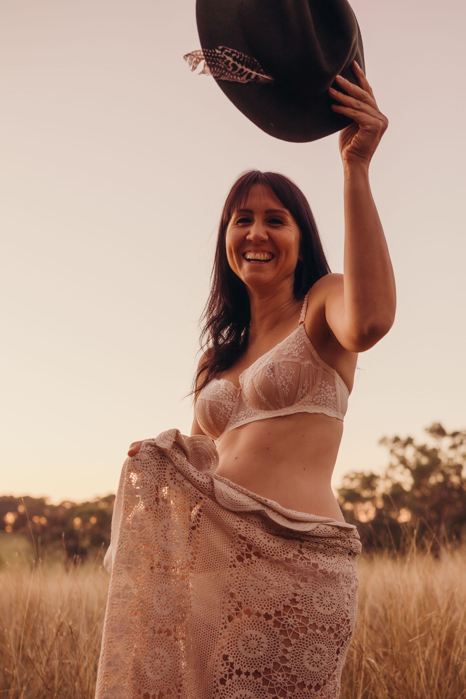 Smiling woman in lace lingerie holding a cowboy hat aloft in a golden field at sunset.