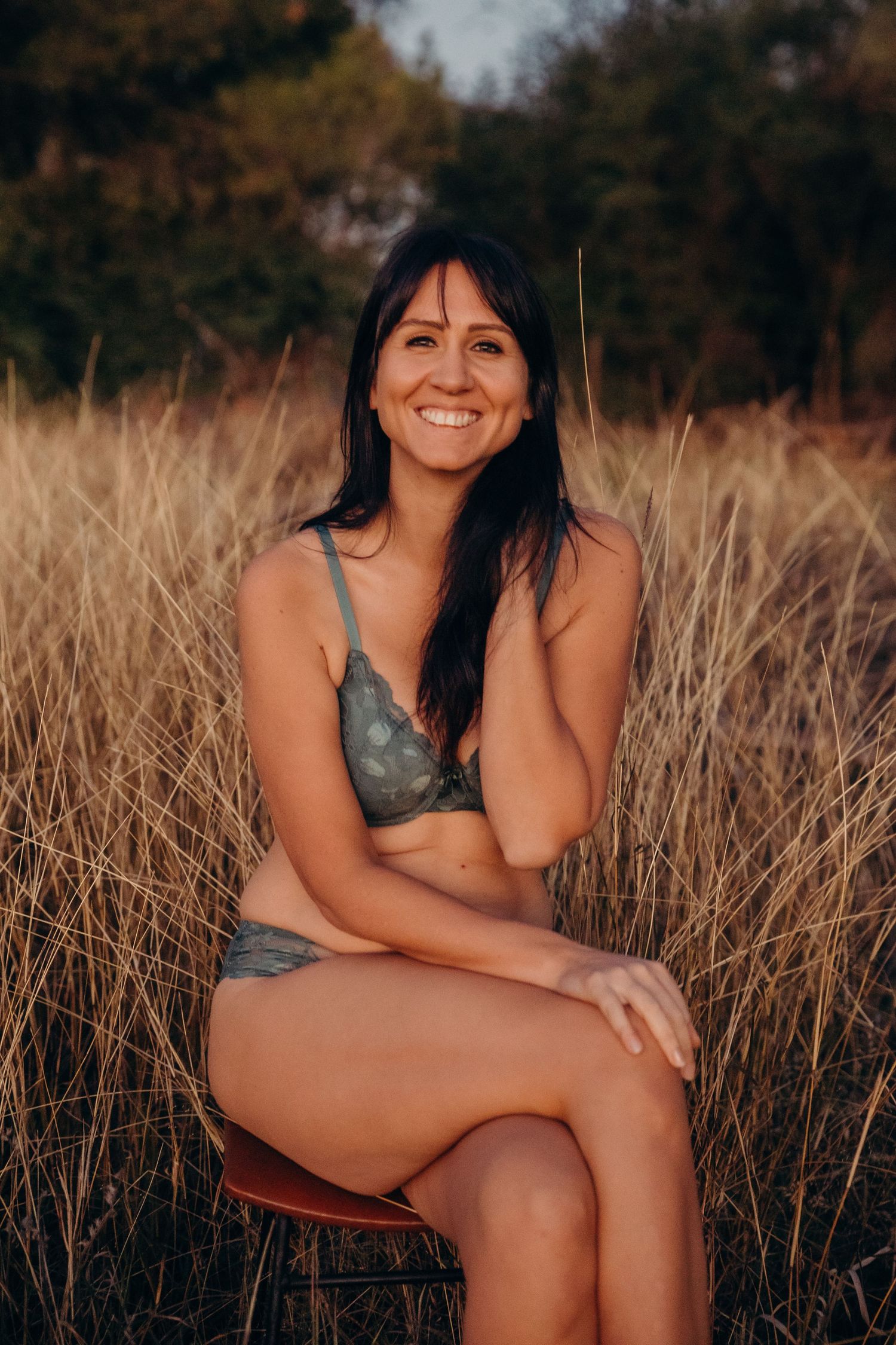 Happy brunette woman in olive green lingerie set sitting in tall golden grass at dusk.