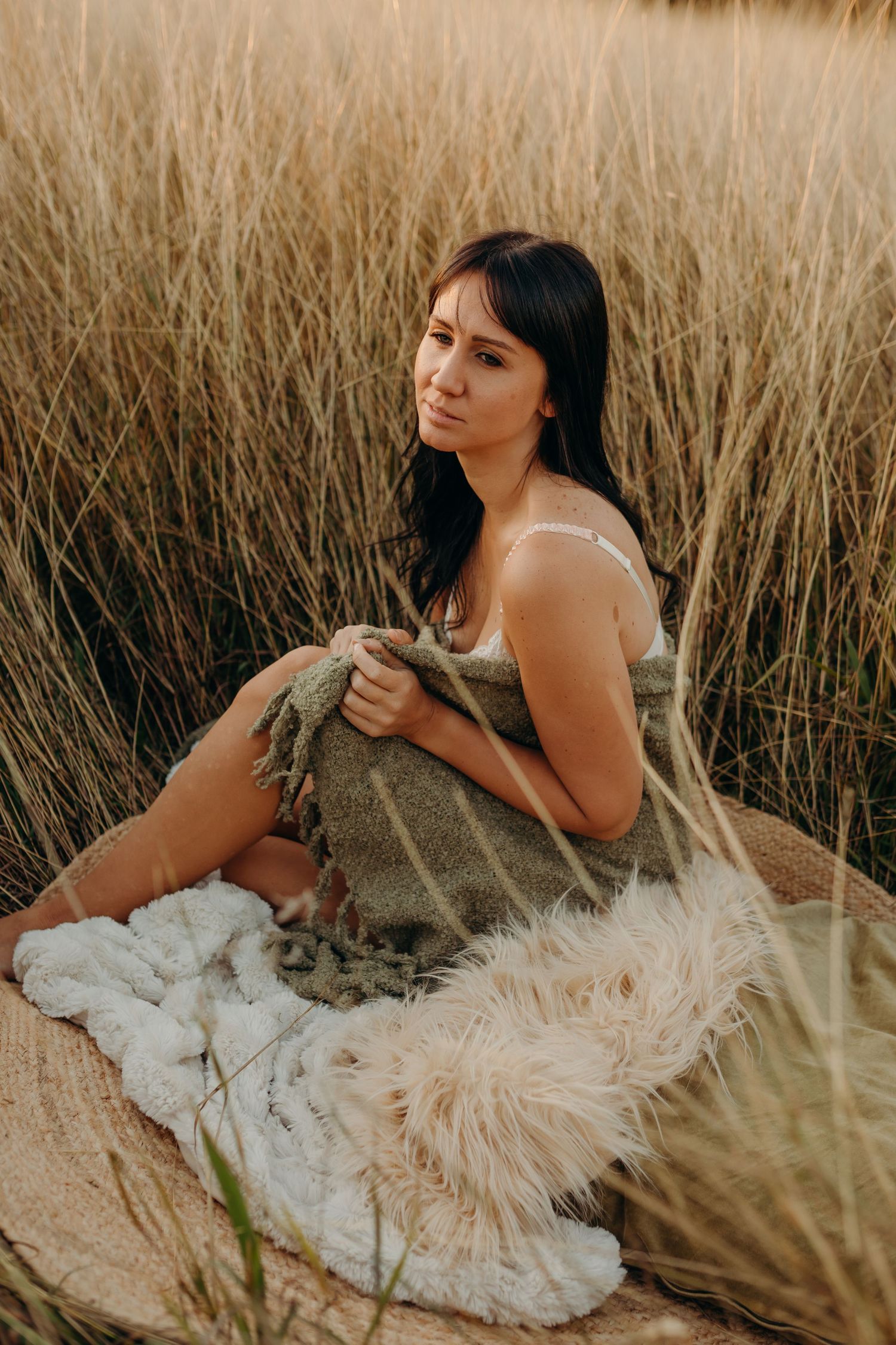 Brunette woman wrapped in a green blanket sitting peacefully in tall golden grass outdoors.