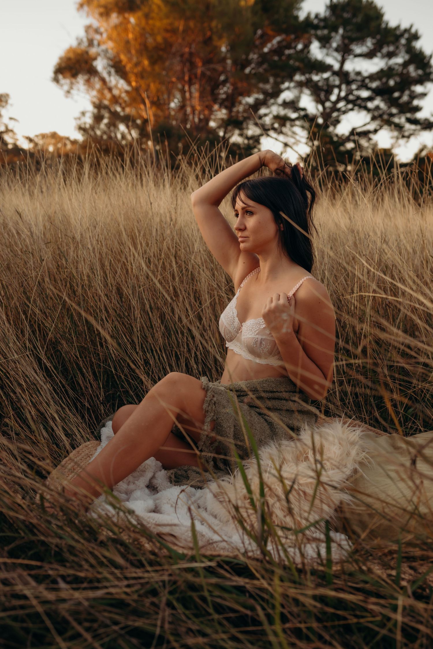 Woman in white lace lingerie posing thoughtfully in tall dry grass at golden hour sunset.
