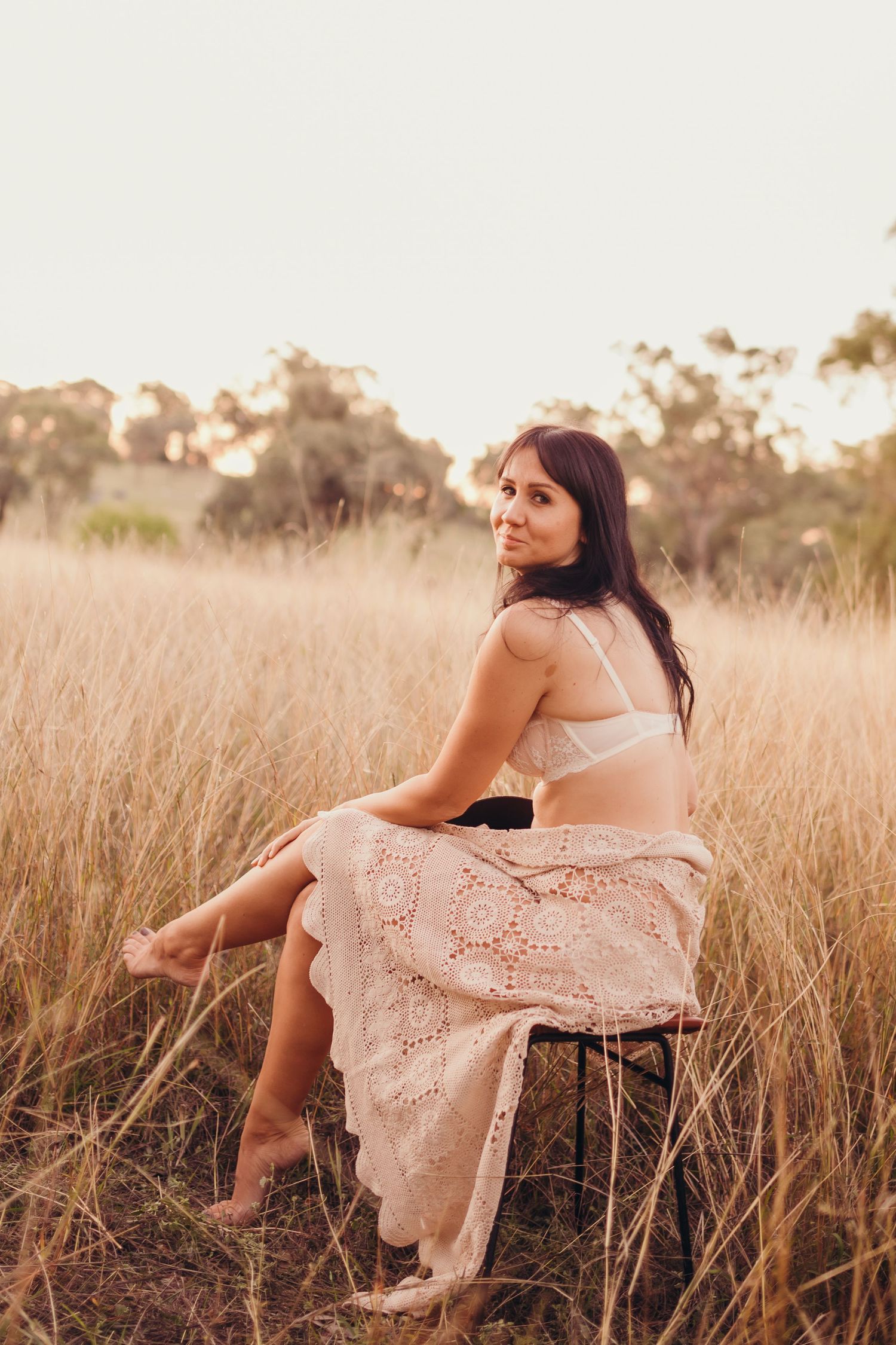 Brunette woman in white lingerie and lace skirt sits on stool in golden grass field.