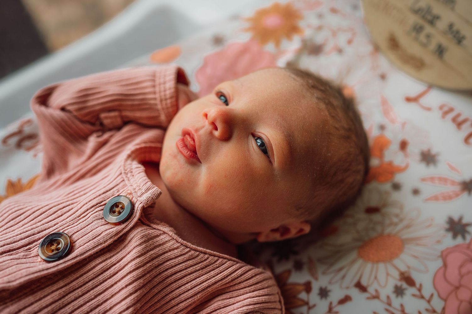 Adorable newborn baby in pink ribbed cardigan lying on floral blanket looking up.