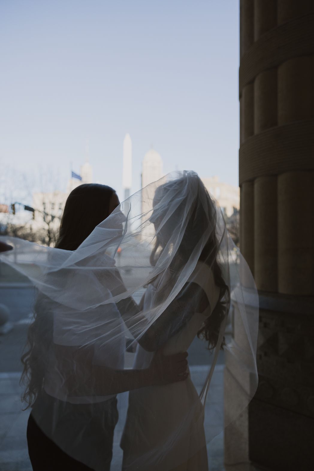 Bride and bridesmaid share a tender moment under a flowing veil with city skyline backdrop.