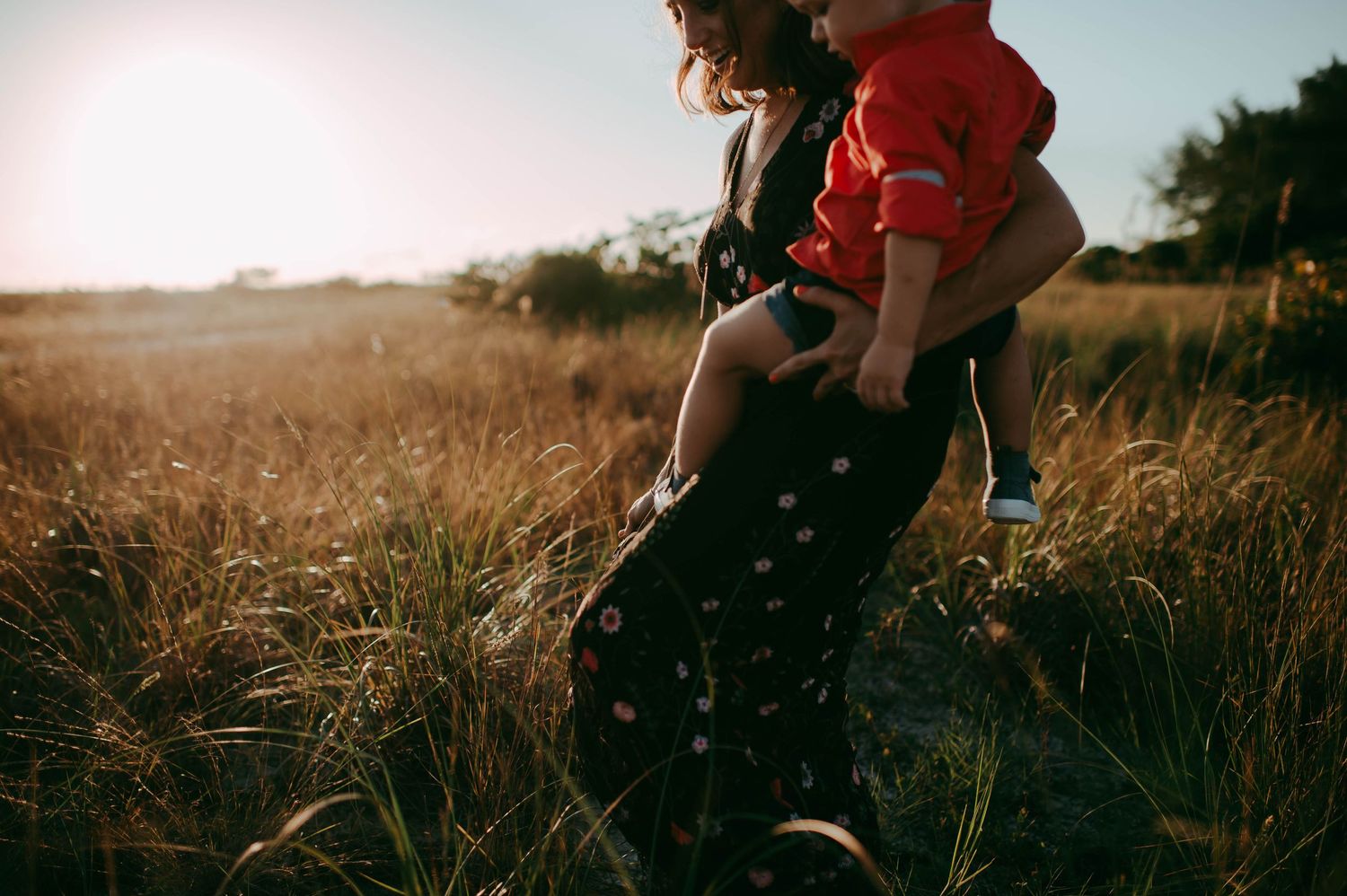 A mother carrying her toddler through a golden sunlit field, wearing a floral dress on a warm sunny day.