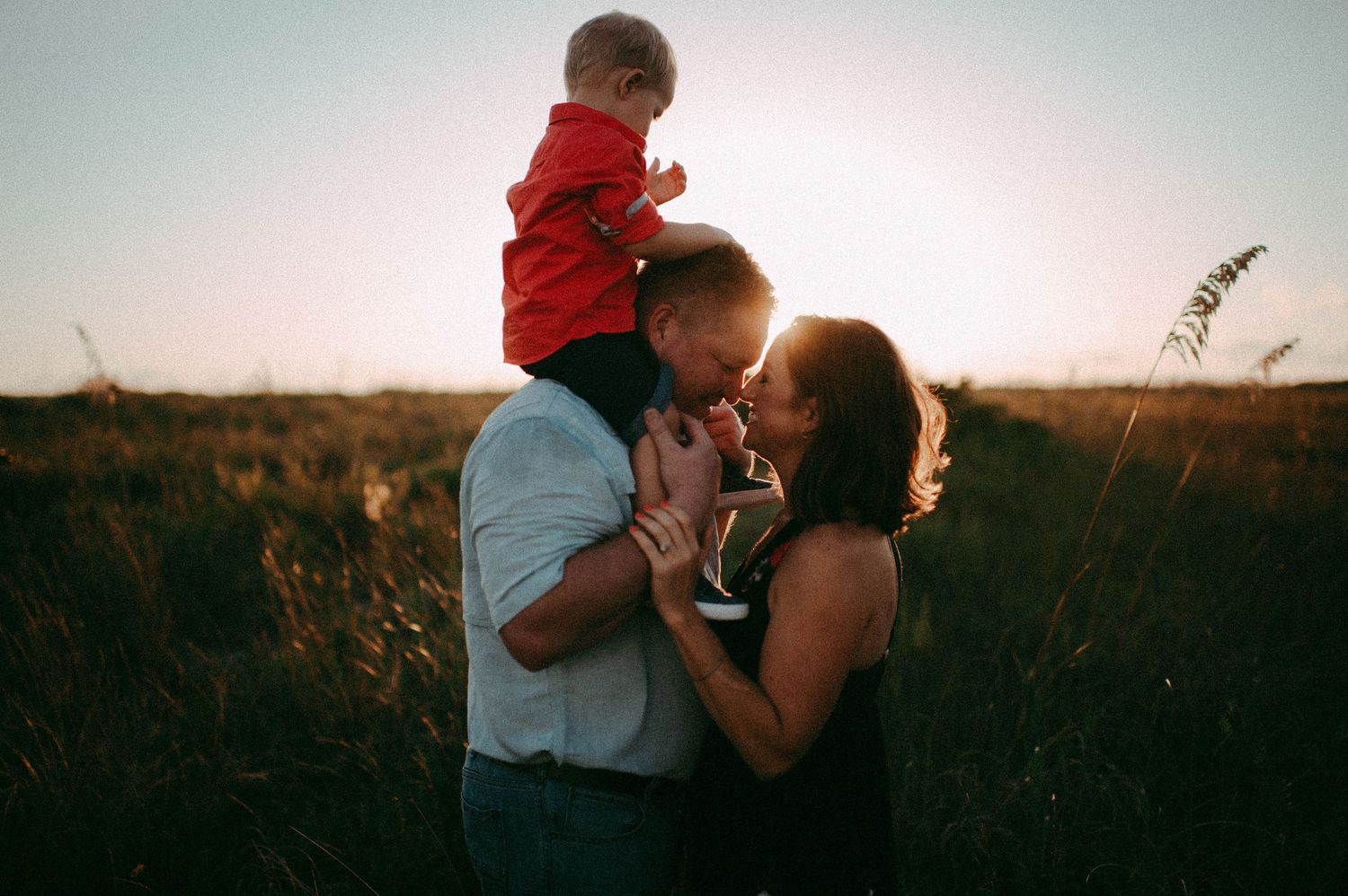 A couple shares a romantic moment in a golden field at sunset while their toddler sits on dad's shoulders.