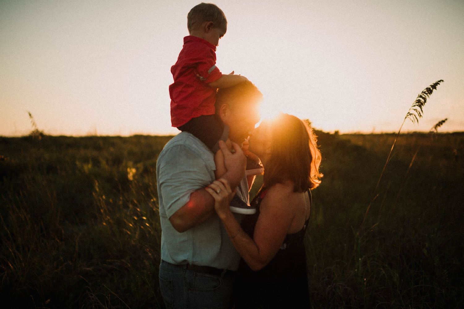 A couple shares a sunset kiss in a field while their young child sits on the father's shoulders.