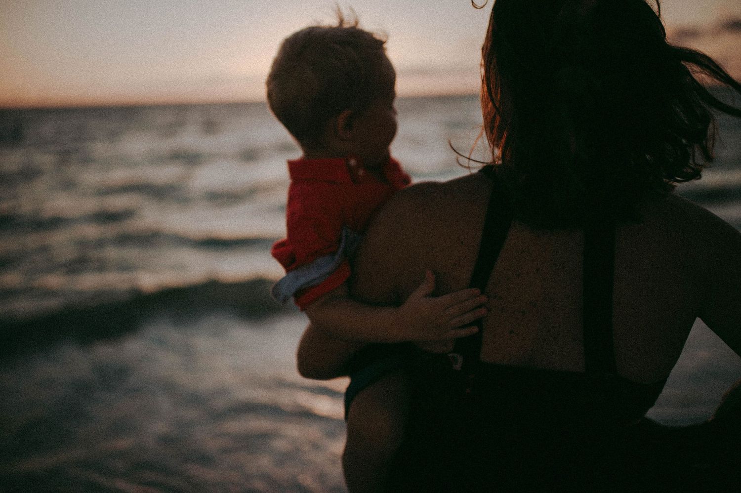 Mother holding young child on beach at sunset, ocean waves in background, warm golden light.