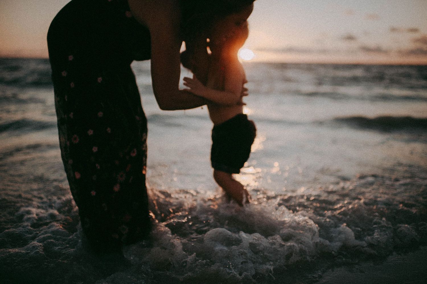 A silhouette of a mother holding a toddler at sunset on a beach with ocean waves.