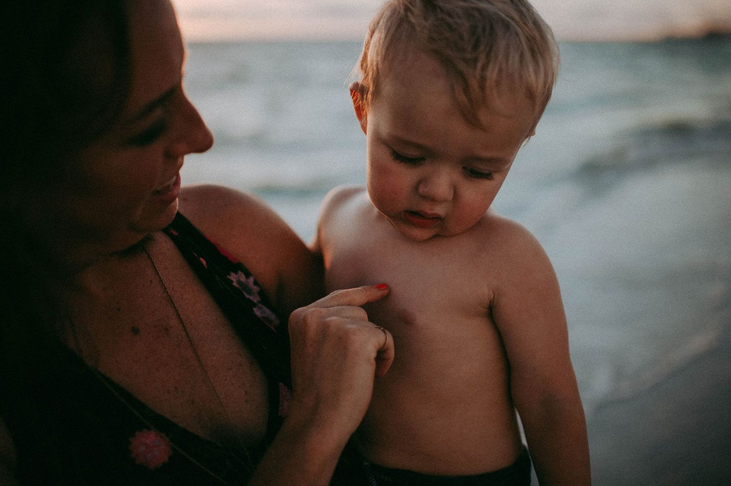 Mother holding toddler boy on beach at sunset, checking skin for sunburn near ocean shoreline.