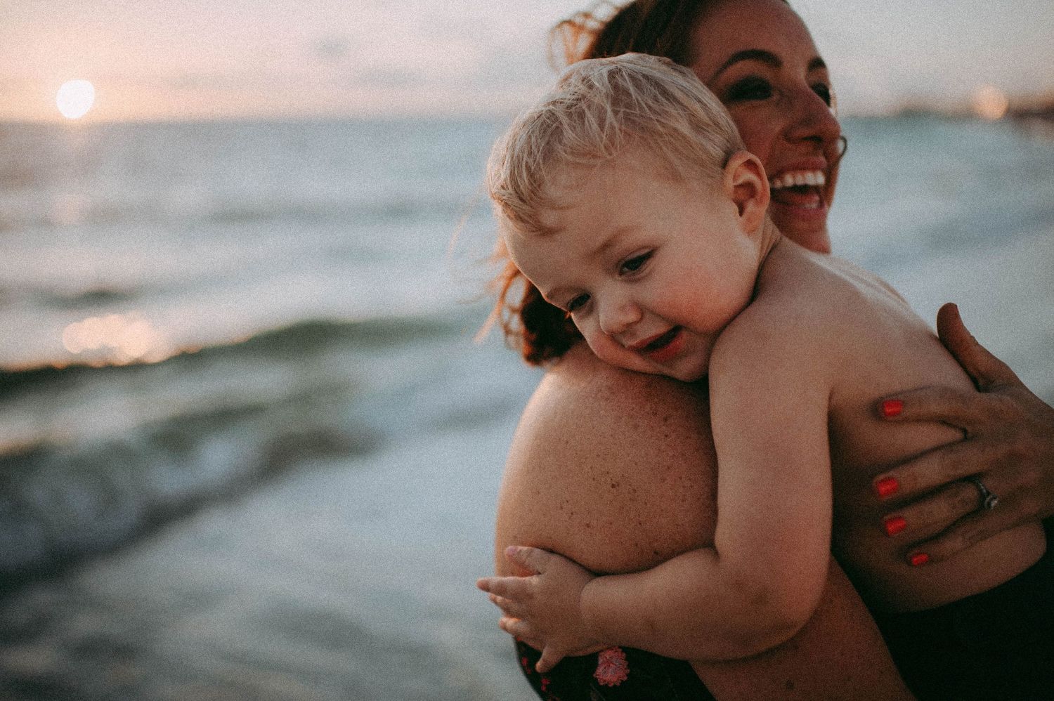 A smiling mother holds her happy toddler on a beach at sunset, both enjoying a joyful moment by the ocean.