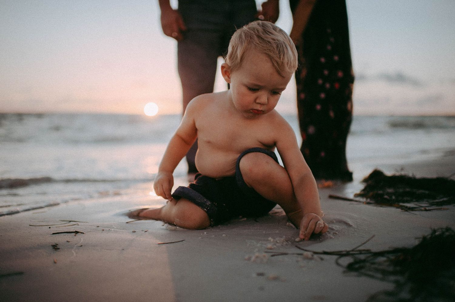 Toddler boy playing in sand at sunset beach with adults standing behind him.