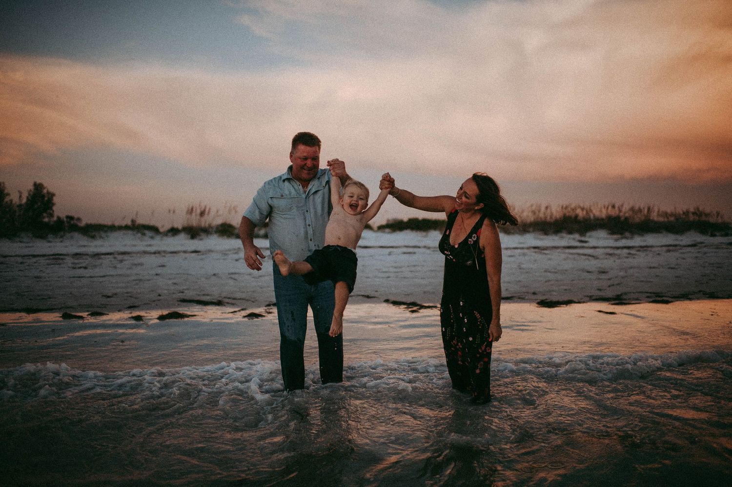 Family of three playing in shallow ocean waves at sunset, parents swinging child between them on sandy beach.