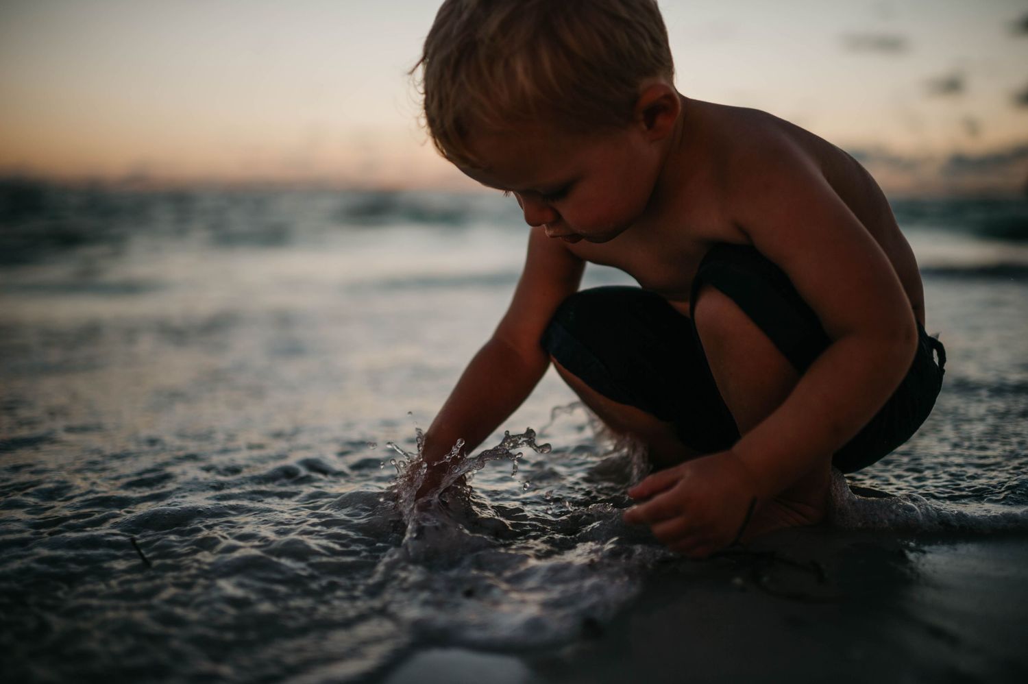 Young toddler boy playing in ocean waves at sunset on a sandy beach.