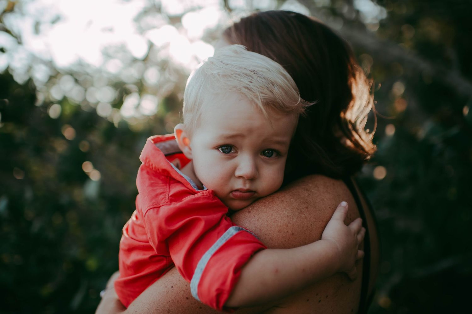 A blonde toddler in a red jacket cuddles closely with a woman, resting his head on her shoulder outdoors.