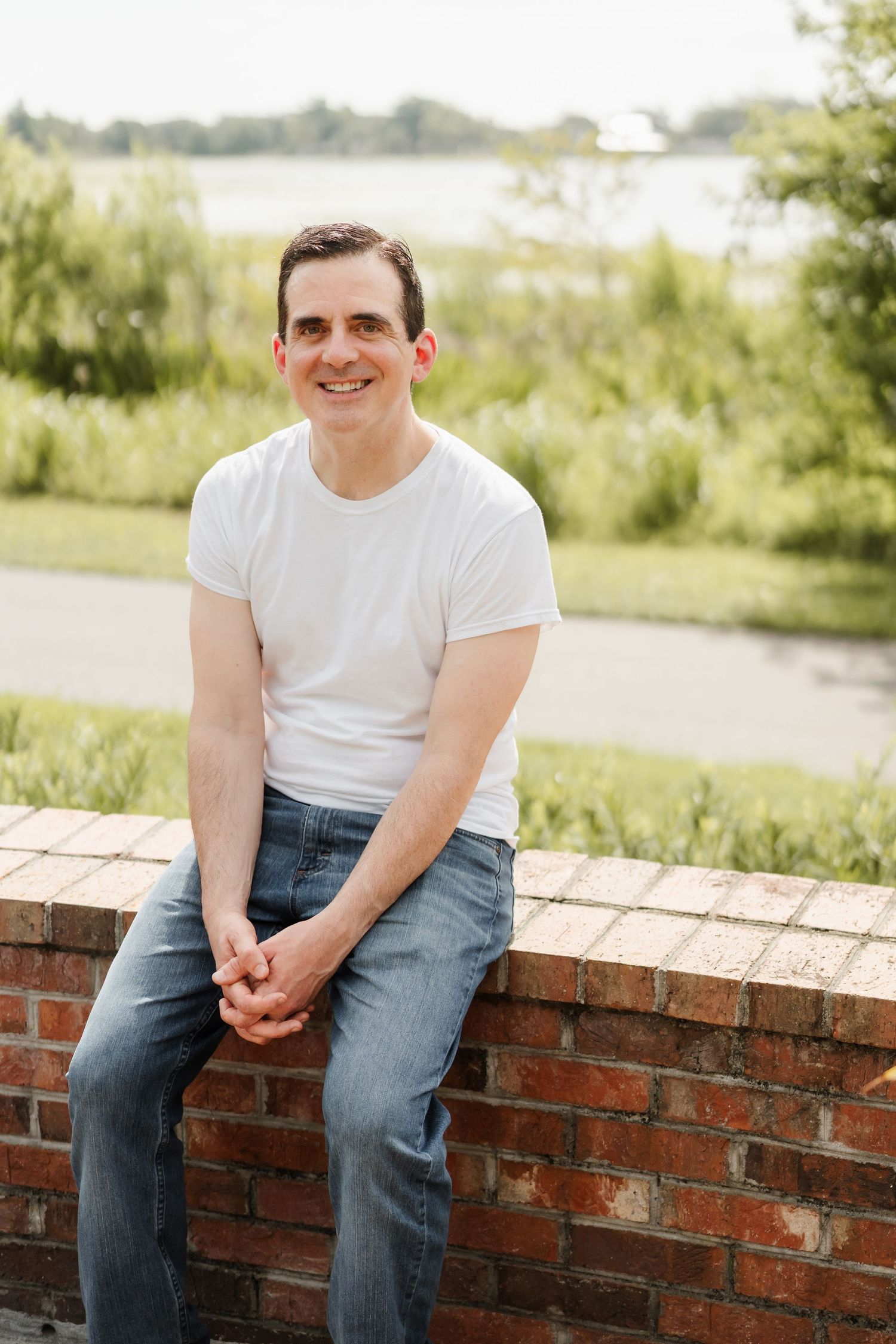 Smiling man in white t-shirt and jeans sitting on brick wall outdoors near a lake with green trees.