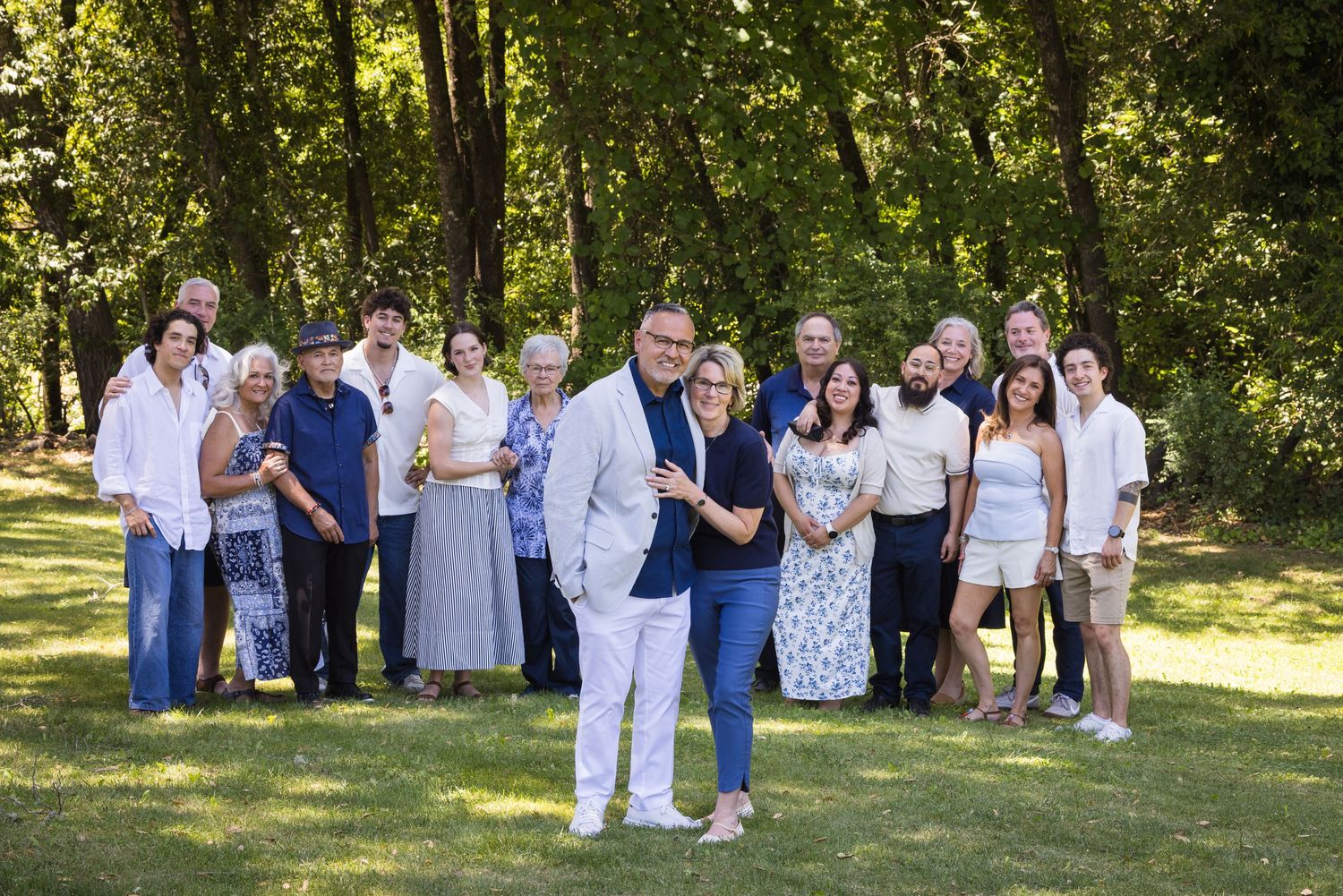 Large family group portrait outdoors in lush green park setting, smiling and posing together on sunny day.
