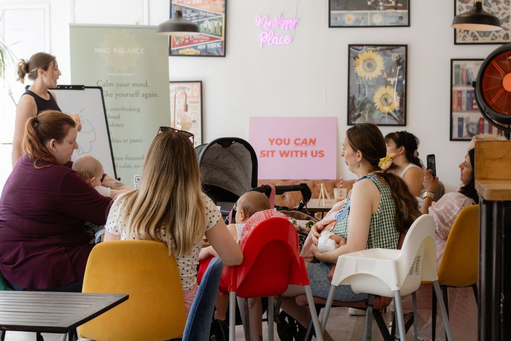 Women gather at a cozy cafe workshop with a 'You Can Sit With Us' neon sign while a presenter leads the group.
