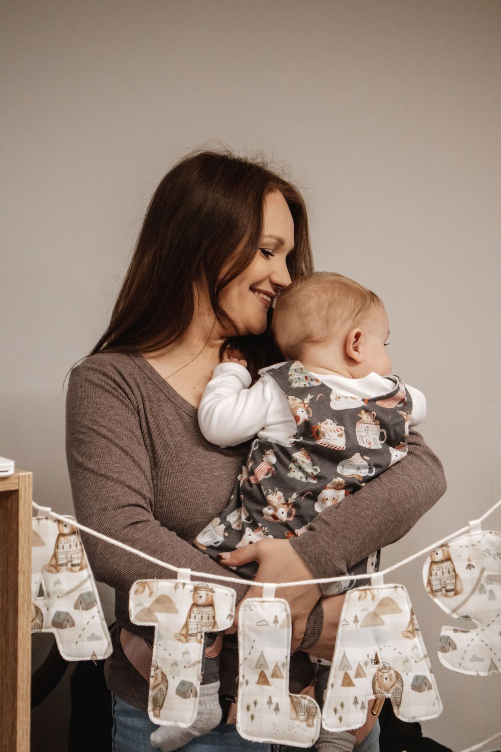 A mother with long brown hair smiles tenderly at her baby while holding them, with a decorative letter banner in the background.