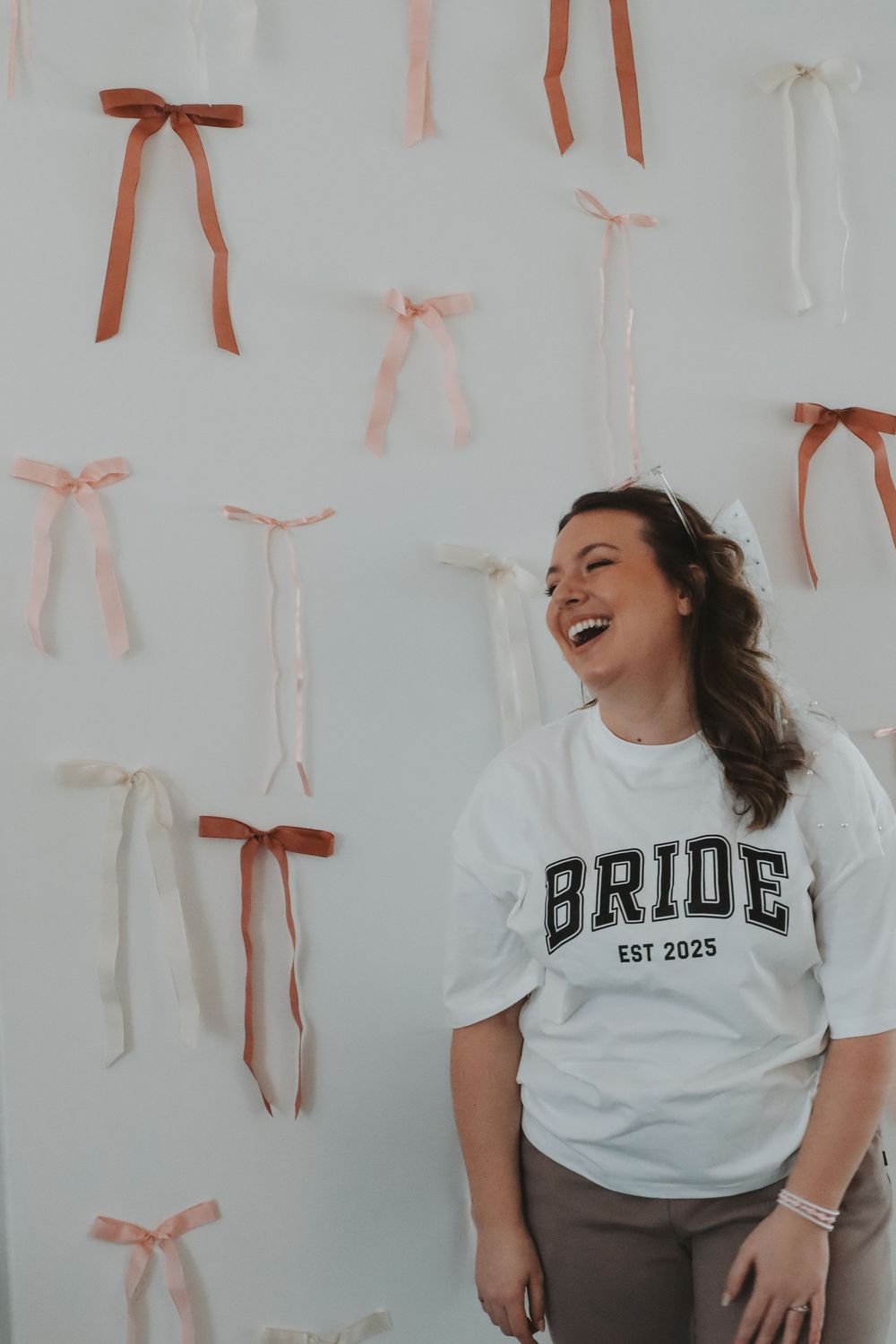 Laughing bride wearing white EST 2025 shirt poses in front of wall decorated with colorful ribbon bows.
