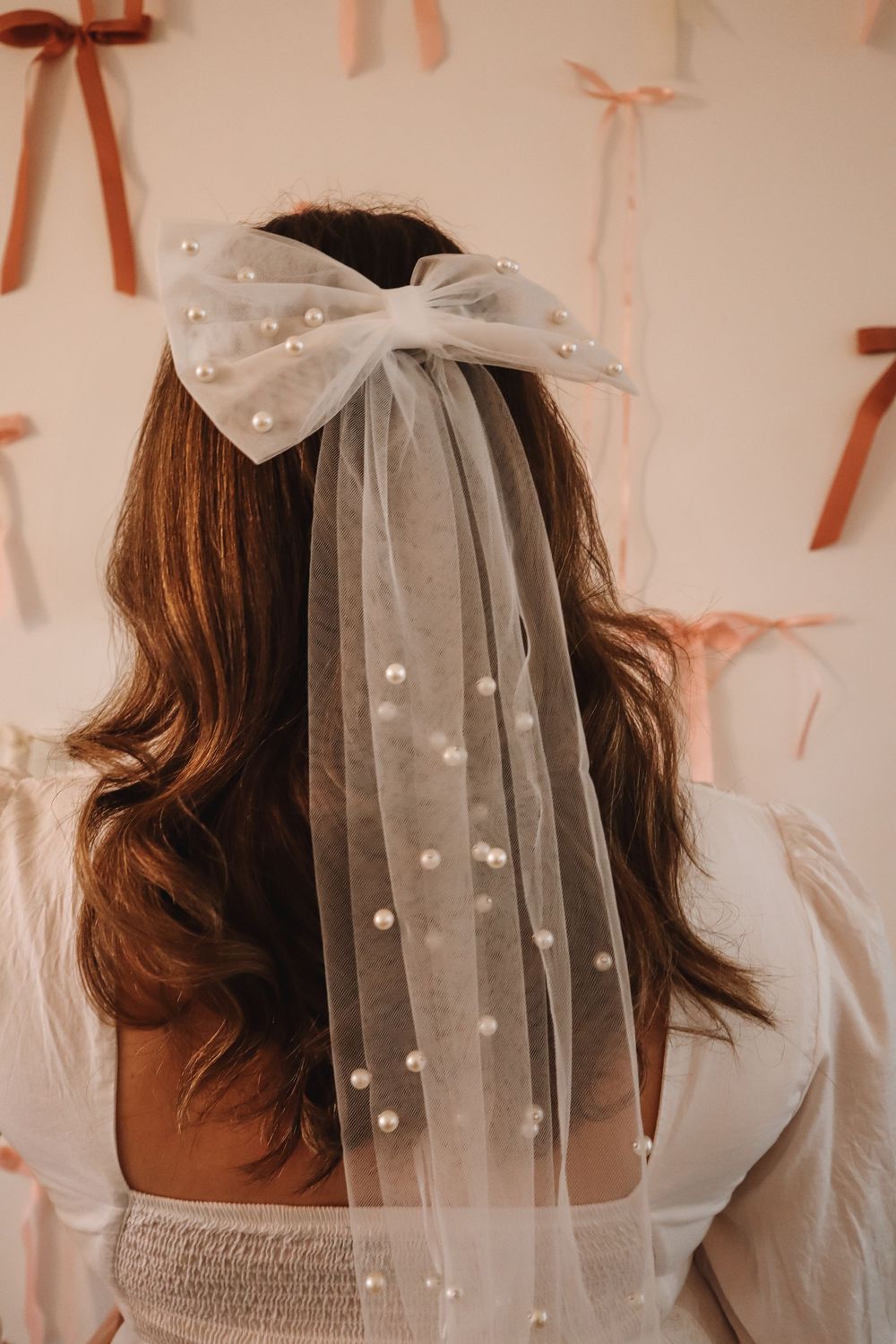 Bridal tulle bow veil with pearl embellishments worn by brunette bride, against pink ribbon wall backdrop.
