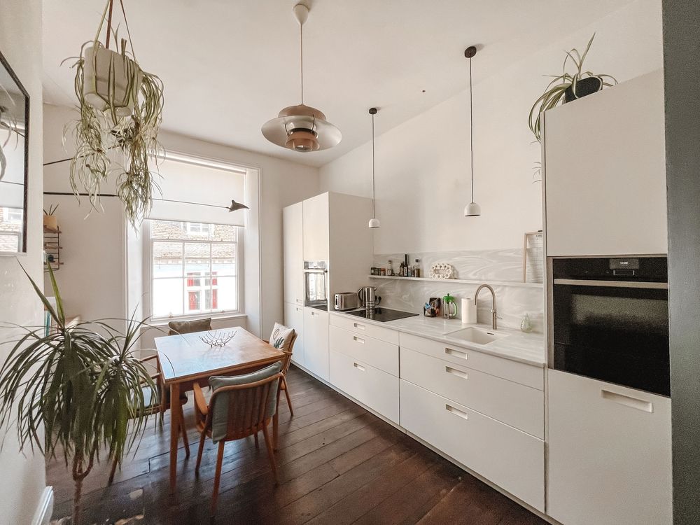 Bright white kitchen with dark hardwood floors, dining table, pendant lights, and lush green plants near a large window.