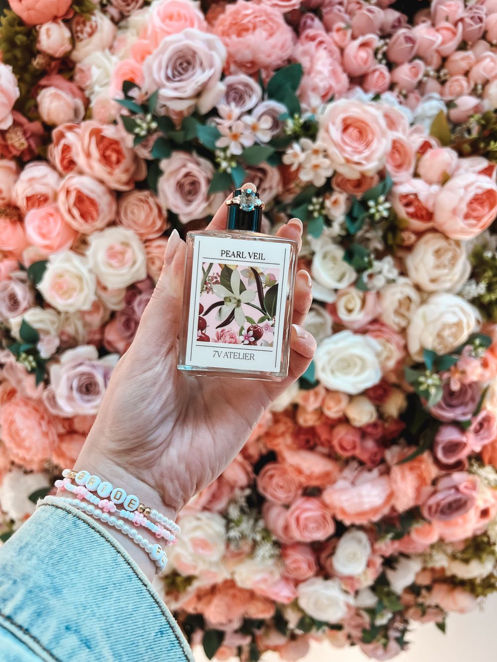 Hand holding Pearl Veil 79 Atelier perfume bottle against a lush wall of pink and peach roses.