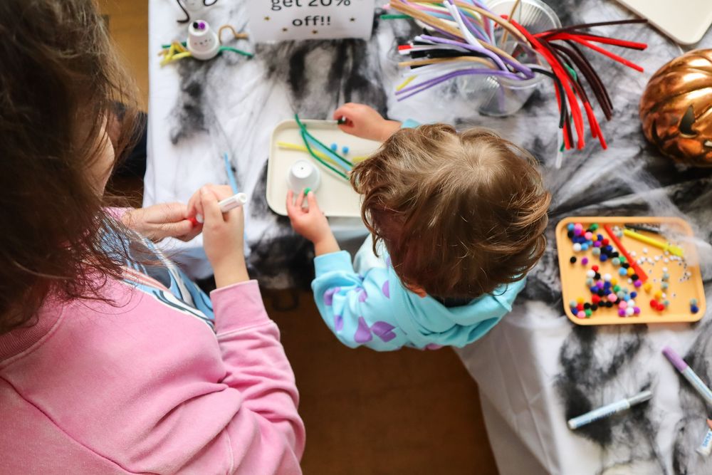 Two children crafting with colorful supplies on a table decorated with Halloween spider web decorations.