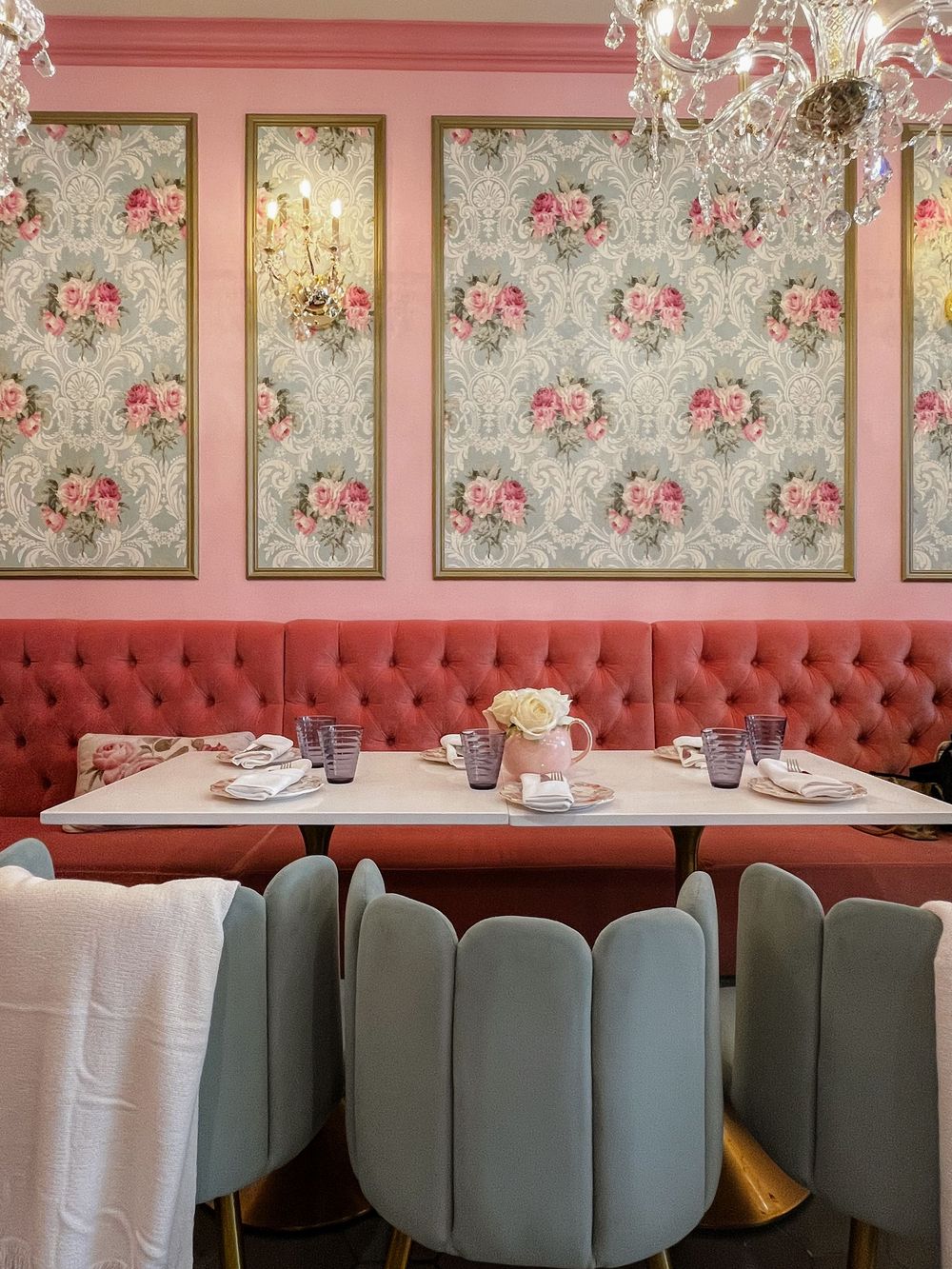 Elegant pink restaurant booth with tufted velvet seating, floral wallpaper panels, marble tables, and sage green chairs.